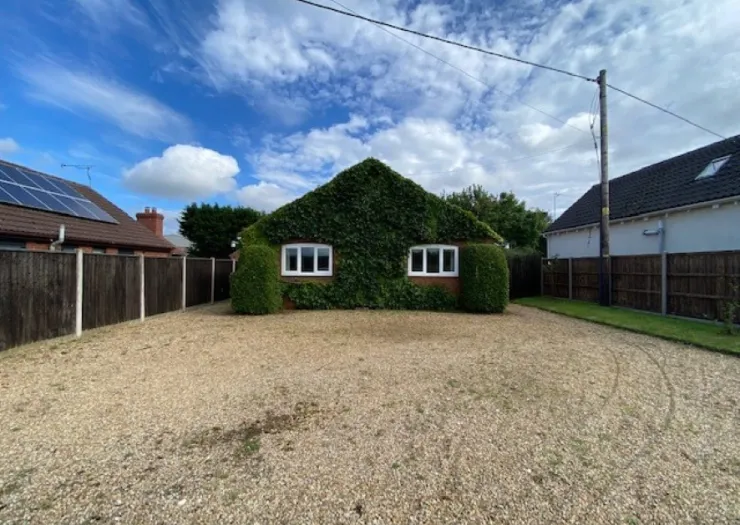 Ivy-covered bungalow with gravel driveway and fenced yard under a blue sky.
