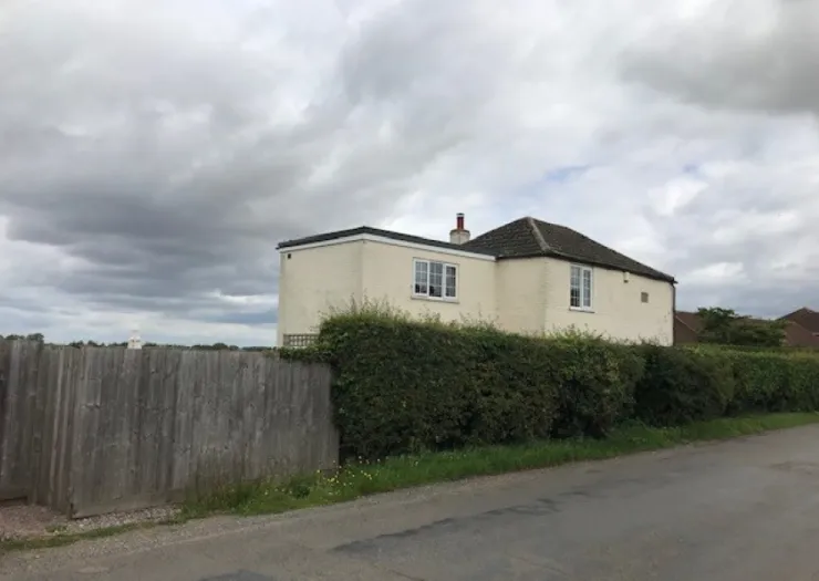 Cream-colored house with a tiled roof, behind a tall hedge and wooden fence, under a cloudy sky.