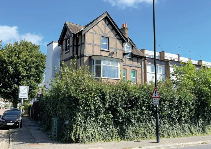 Tudor-style house with bay window behind a tall hedge. Road works sign visible.