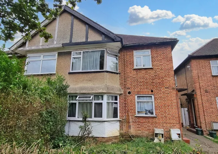 Exterior view of a semi-detached brick house with bay windows, overgrown front garden, under a blue sky.