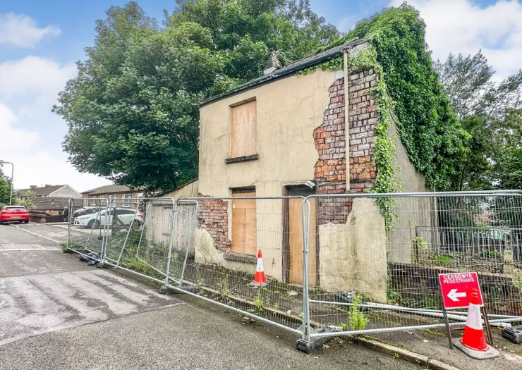 Derelict building with boarded windows, crumbling facade, and metal fencing. Pedestrian detour sign visible.