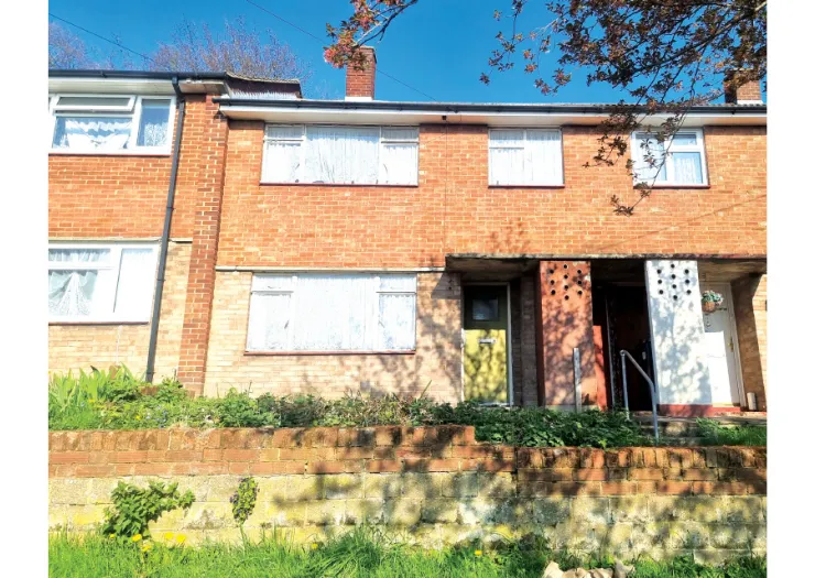 Attached brick house exterior with white-curtained windows, a low brick wall, and green front lawn.