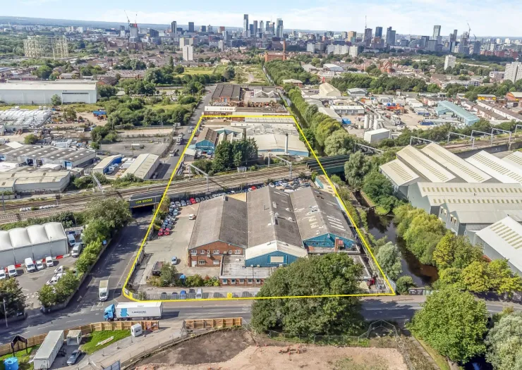 Aerial view of industrial buildings outlined in yellow, near a canal and railway, with a city skyline in the background.