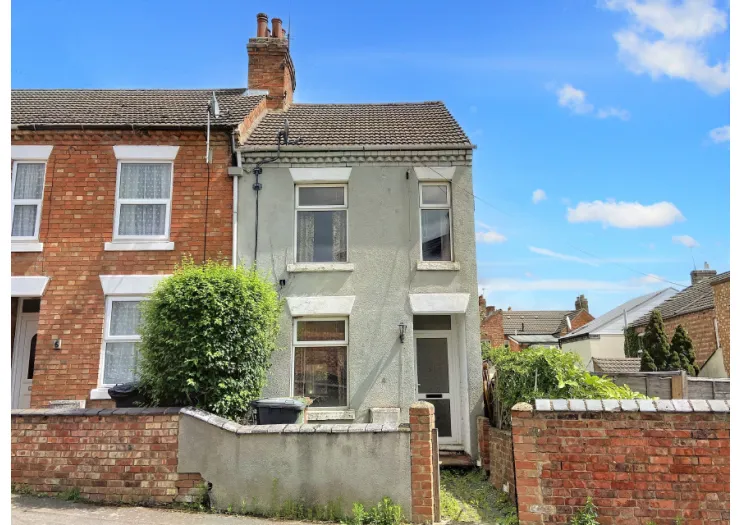 Two-story terraced house with gray facade, white door, and brick garden wall under a blue sky.