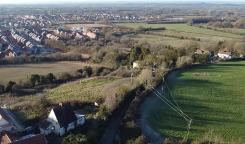 Aerial view of a rural landscape with houses, fields, and a new housing development in the distance.