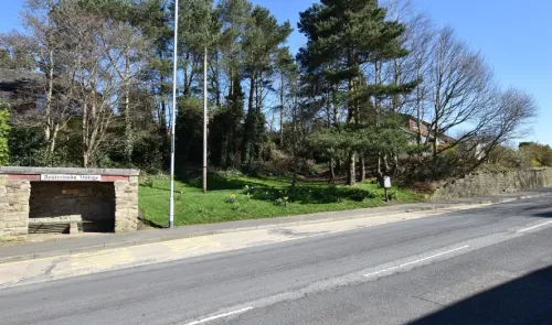 Austerlands Village bus stop with grassy area, trees, and stone wall on a sunny day.
