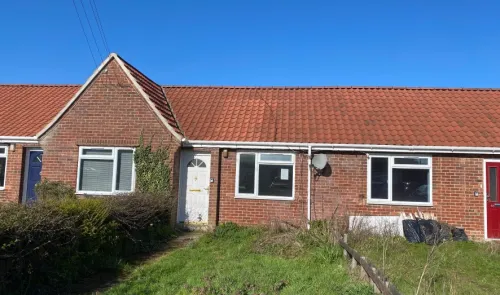 Brick bungalow with red tiled roof, white-framed windows, and overgrown front garden.