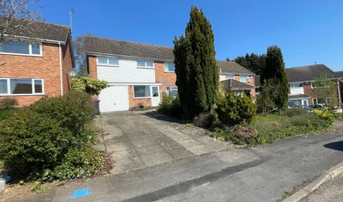 Suburban house exterior with driveway, front garden, and tall evergreen trees.
