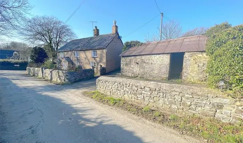 Charming rural cottage with attached stone barn and low stone wall along a quiet country lane.