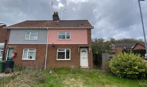 Pink and grey semi-detached house with front garden and overgrown bushes.