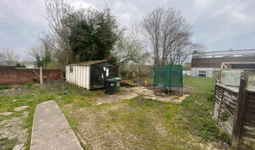 Overgrown garden with shed, trampoline, wheelie bins, and concrete path.
