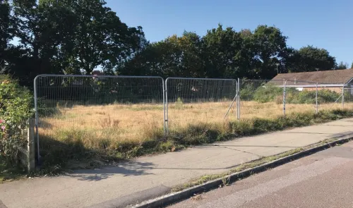 Empty fenced lot with overgrown grass, sidewalk, and trees.