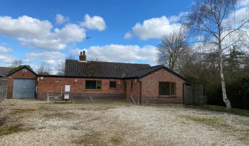 Red brick bungalow with attached garage, gravel driveway, and trees under a blue sky with fluffy clouds.