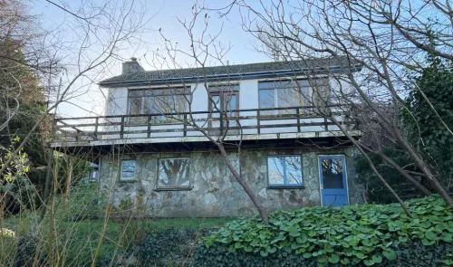 Two-story house with balcony, stone lower level, and surrounded by trees and greenery.