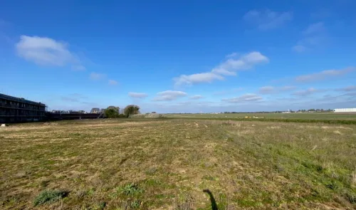 Empty field with sparse grass, a building under construction in the background, and a clear blue sky.