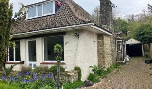 Suburban house with tiled roof, stone chimney, and brick driveway. Front garden with purple flowers. Needs renovation.