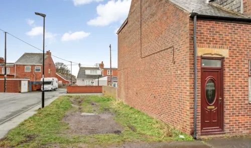 Brick house exterior with burgundy door beside a grassy patch and street view of other houses.