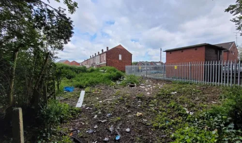 Overgrown vacant lot with litter, fenced area, and brick buildings in the background.