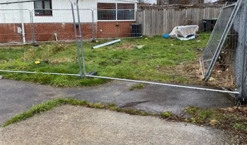 Overgrown grassy area behind metal fencing, with debris and a partially collapsed fence section.