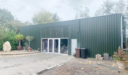 Green metal industrial building with glass doors, viewed from paved driveway. Landscaping and potted plants in front.