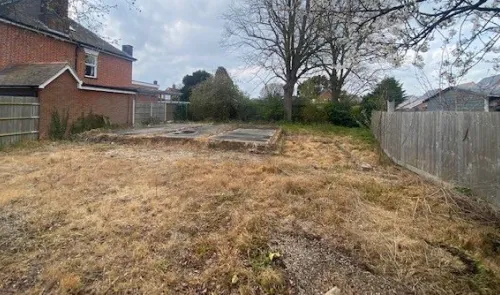 Overgrown lot with concrete foundation remnants, trees, and a partial view of a brick house.