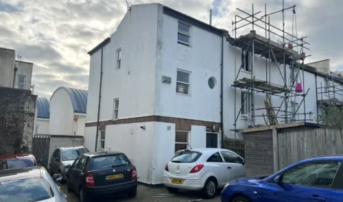 White building with scaffolding, small circular window, and parked cars in front.