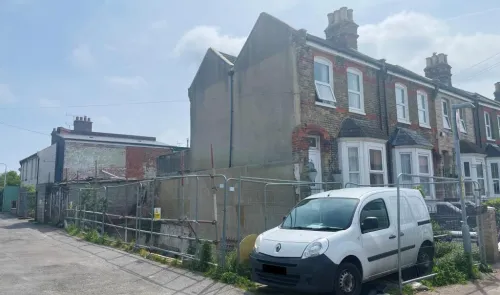 Row of terraced houses, one with exposed side wall and construction fencing. White van parked on street.