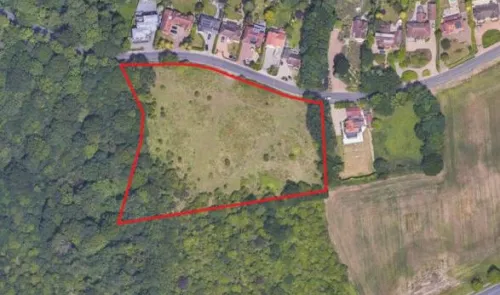 Aerial view of vacant land outlined in red, bordering a road and houses, surrounded by trees.