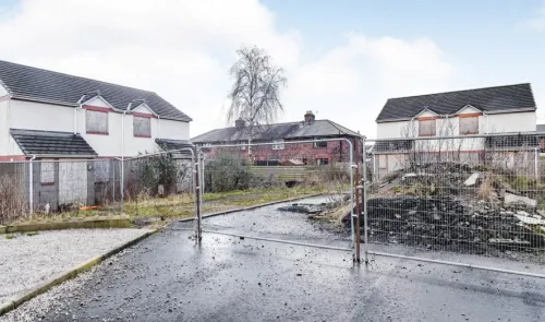Boarded-up houses, demolition rubble, and metal fencing on a residential street.