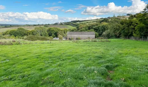 Green field with a barn, trees, and rolling hills under a blue sky with fluffy white clouds.