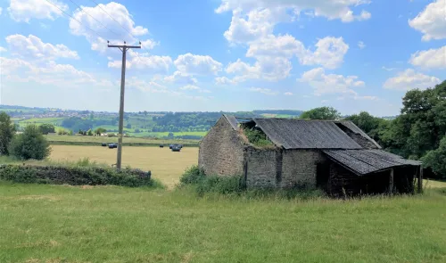 Dilapidated stone barn in a green field, with hay bales and rolling hills in the distance under a blue sky.