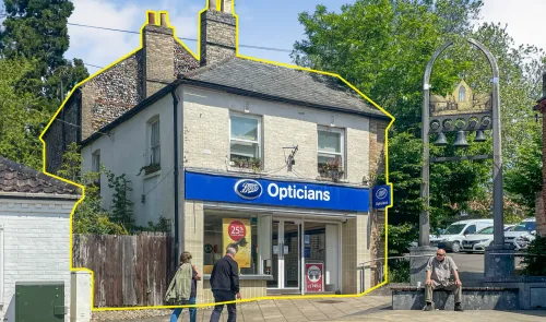 Boots" Opticians storefront in a two-story building, pedestrians passing by, village bell structure visible.