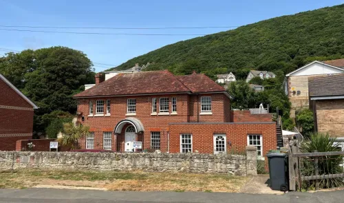 Red brick house with arched entryway, stone wall, and forested hillside backdrop.
