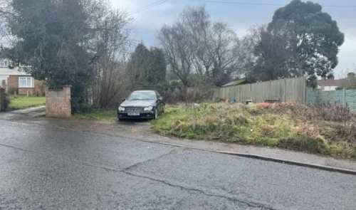 Overgrown plot of land beside a road with a parked car, trees, and fences.
