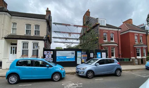 House under construction between two existing terraced houses, supported by scaffolding. Construction site signage visible.