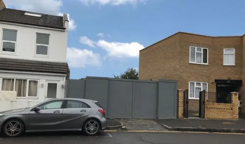 Gray hoarding between houses, parked car, and open metal gate.