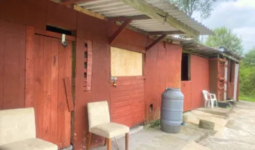 Red wooden outbuildings with boarded-up windows, a water barrel, and chairs outside.