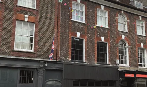 Brick building with dark storefronts and Union Jack flag. Upper levels have multi-paned windows and festive bunting.