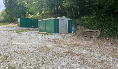 Two green metal storage sheds on gravel area near trees.