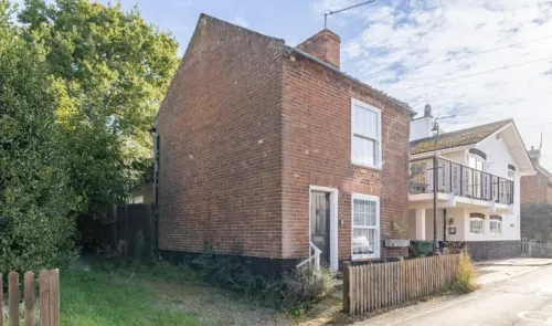 Charming red brick cottage with white trim, wooden fence, and neighboring building with balcony.
