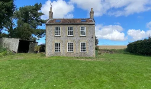 Stone farmhouse with white-framed windows, two chimneys, and a green lawn under a blue sky.