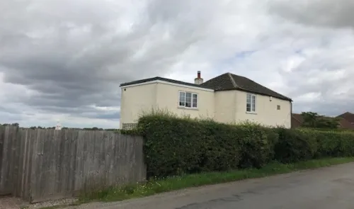 Cream-colored house with a tiled roof, behind a tall hedge and wooden fence, under a cloudy sky.