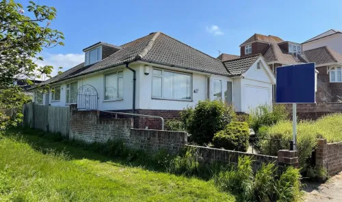 White bungalow with gray tiled roof, front garden, and "For Sale" sign.