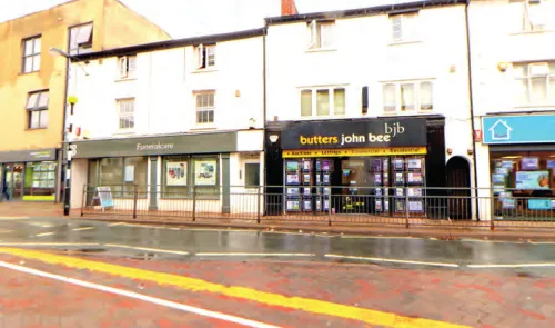 Butters John Bee estate agents storefront with property listings displayed. Adjacent is Funeralcare. Wet street.