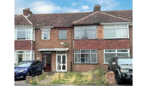 Attached brick house exterior with bay windows, overgrown front garden, and parked cars.